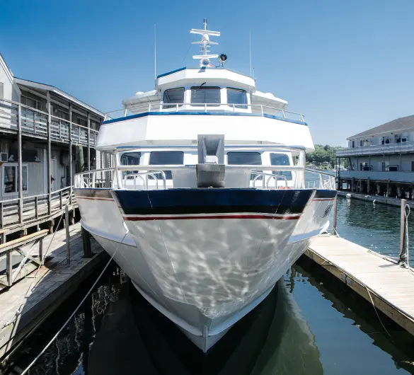 A large white and blue boat is docked in calm water, seen from its bow. The boat has multiple decks and windows, with a blue stripe along its hull. Wooden docks and buildings line both sides of the water under a clear blue sky.
