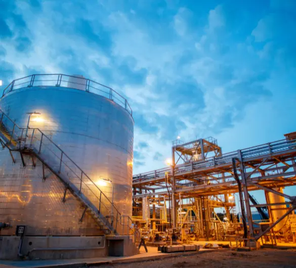 An industrial facility at dusk, featuring a large cylindrical storage tank with stairs leading up its side on the left, illuminated by lights. In the background, there are complex structures of pipes and scaffolding, also lit up, under a cloudy blue sky.