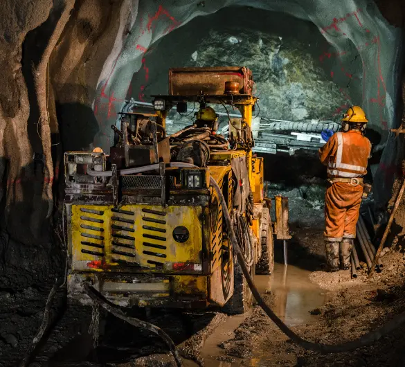 A large, yellow industrial drilling machine is positioned in a dark, wet underground tunnel. A worker in an orange jumpsuit and hard hat stands to the right of the machine, observing the drilling operation. The tunnel walls are rough and rocky.