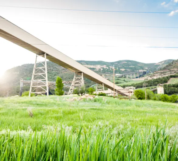A long industrial conveyor belt extends across a vibrant green field, supported by tall metal structures. In the background, hills are visible, and industrial buildings are nestled into the landscape under a bright sky. The foreground features blurred tall green grass.